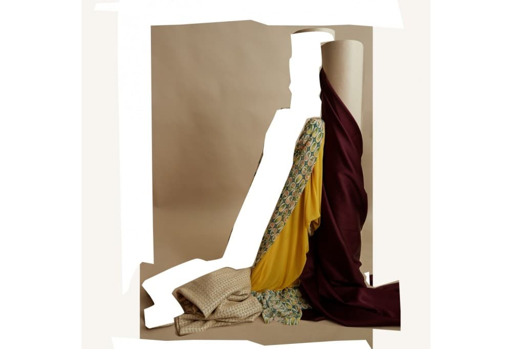 Collage photo : tissus imprimé, jaune et bordeaux drapés sur de grands rouleaux, étoffe beige texturée pliée au sol, devant un fond kraft