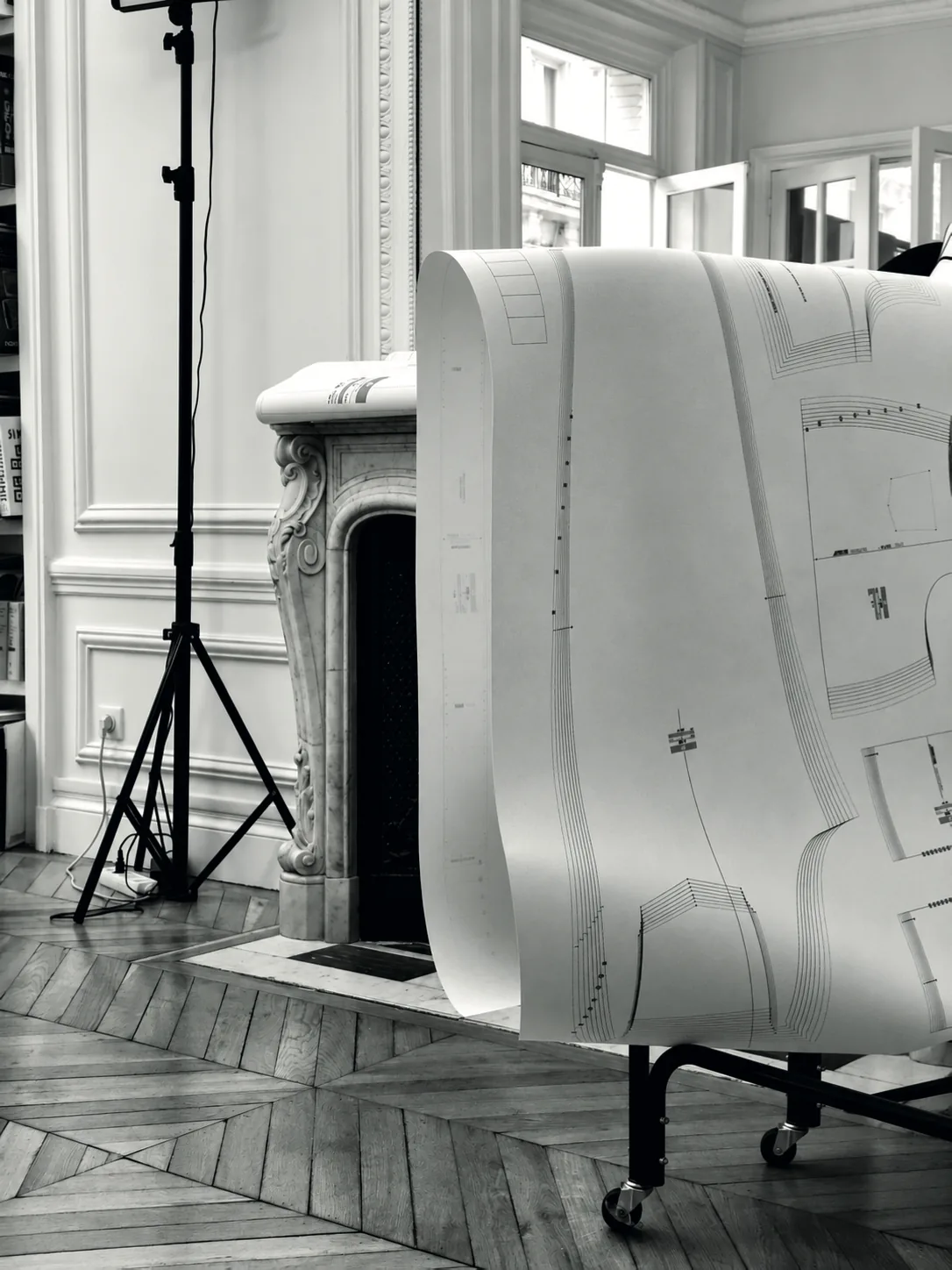 A long printed pattern unrolled over a rail, in front of a fireplace in the studio, in black and white