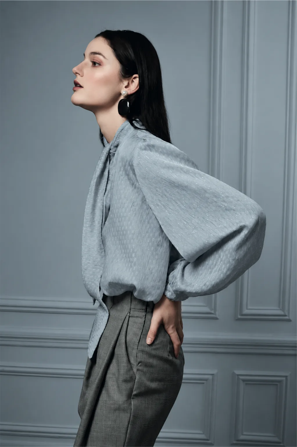 A woman in profile in a blue blouse with full sleeves and checked trousers