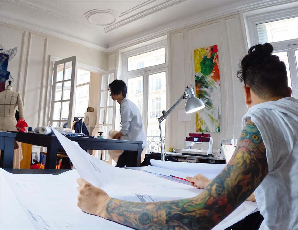 The dp studio atelier in Paris: a patternmaker reading a large pattern, another working in the background, by the windows