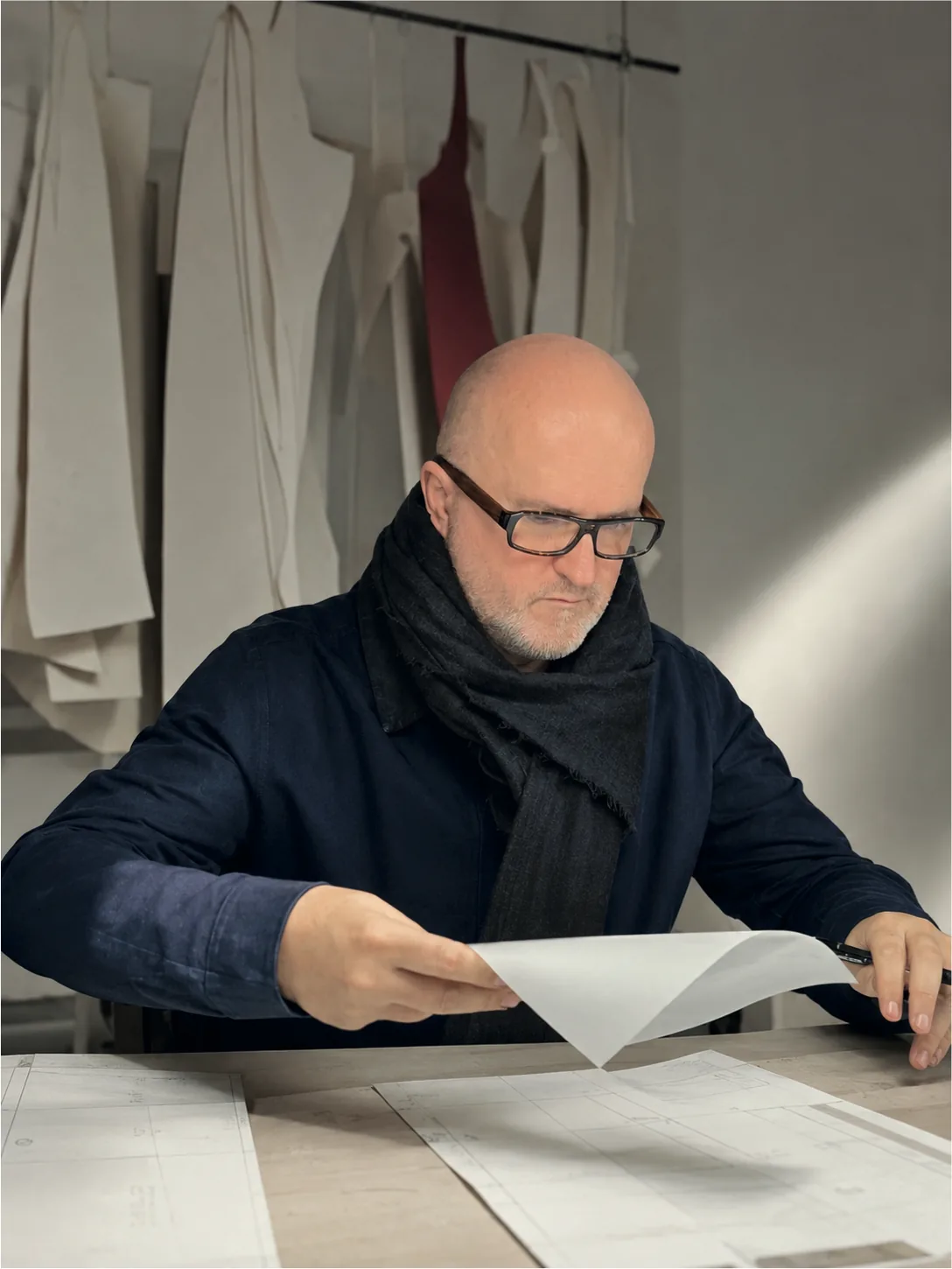 Dominique Pellen in a navy jacket and scarf, tracing paper in hand above his drafts, patterns hanging behind him