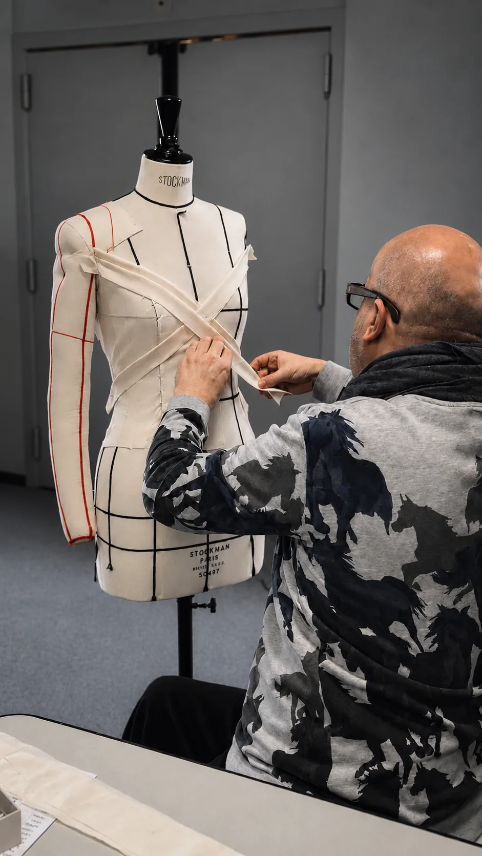 A patternmaker, seen from behind, crosses strips of calico over a Stockman dress form