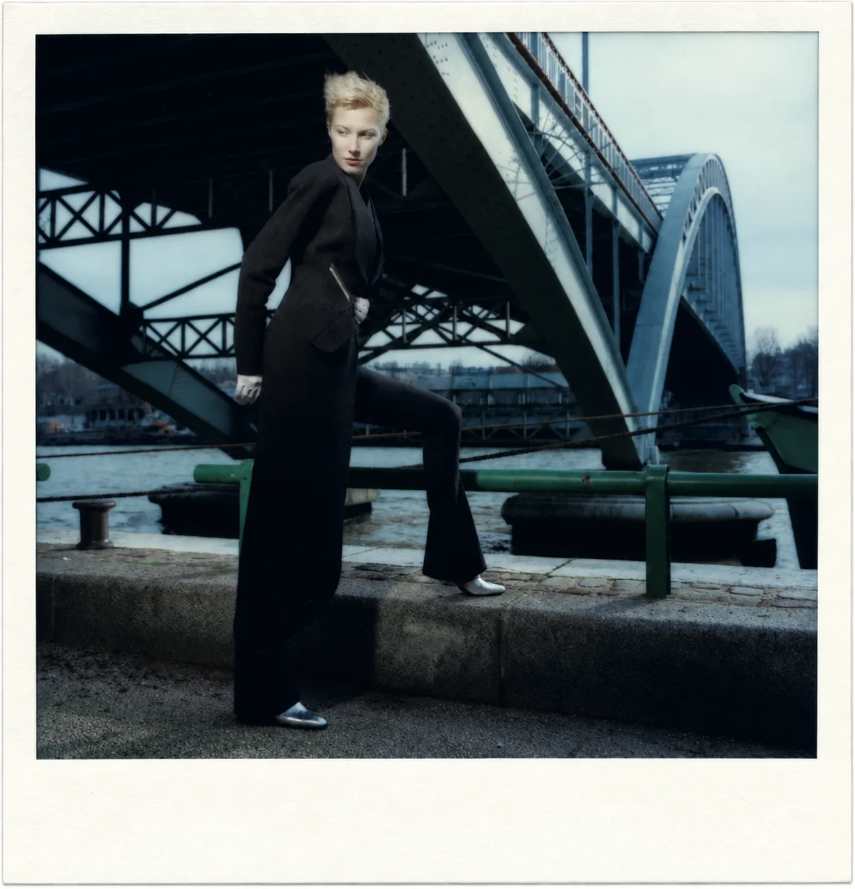 Polaroid: a figure in a black trouser suit, one foot resting on the edge of the quay, beneath the steel frame of a bridge