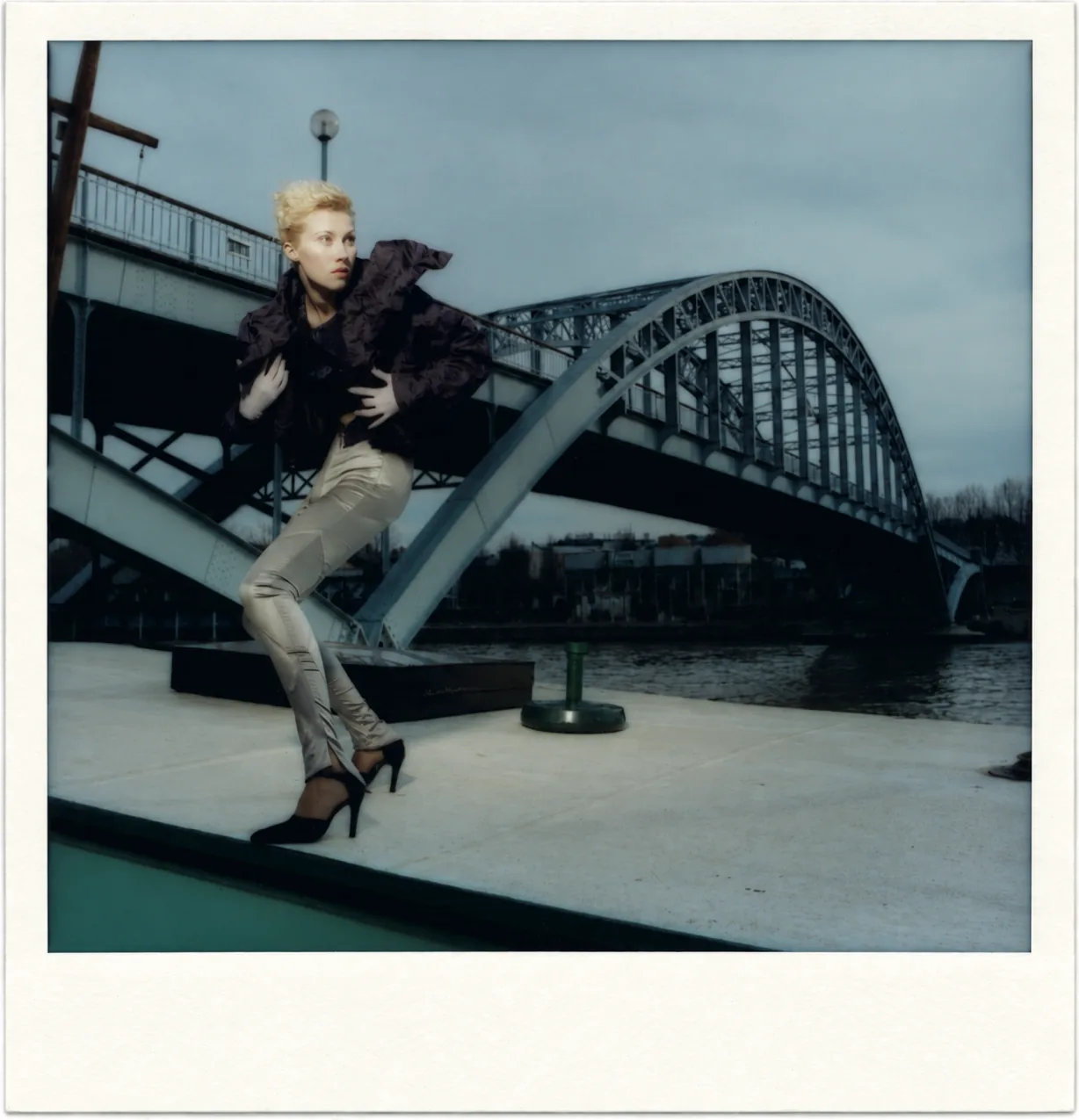 Polaroïd : une silhouette en veste violette à grand col et pantalon argenté, en mouvement sur un quai, devant un pont en arc