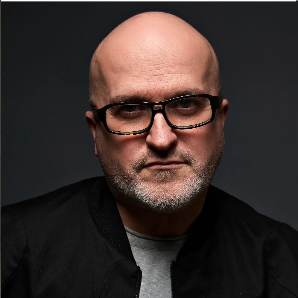 Portrait of Dominique Pellen: shaved head, short grey beard, dark-framed glasses and a black jacket, against a dark background